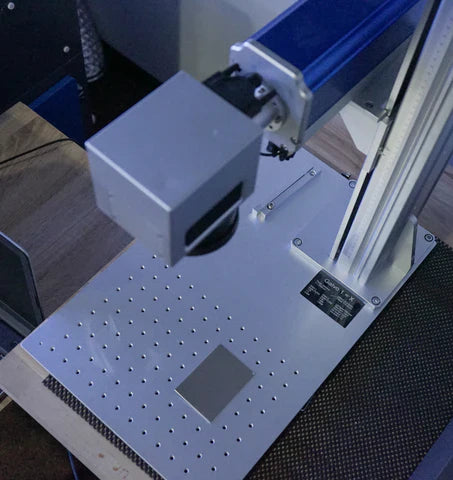 Fiber laser marker head above the worktable with a perforated plate and a metal test piece.
