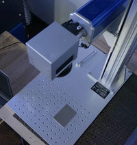 Fiber laser marker head above the worktable with a perforated plate and a metal test piece.