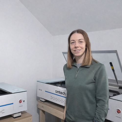 Woman standing next to two Omtech laser cutting machines in a workshop.